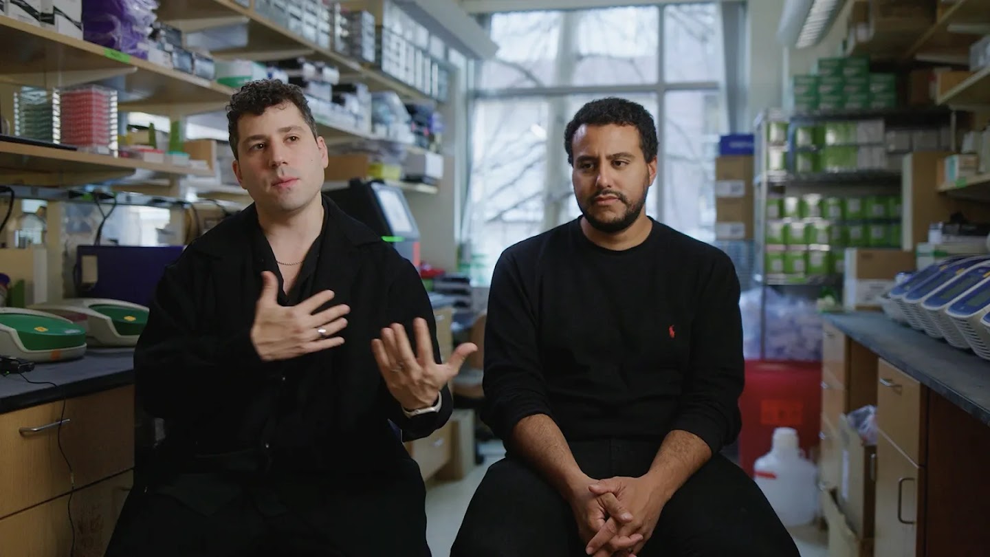 Image 37: This image shows two men sitting side by side in what appears to be a laboratory setting. The man on the left is wearing a black top and has both hands raised, as if speaking or gesturing. The man on the right has a beard and is wearing a black long-sleeve top, looking forward with a serious expression. In the background, shelves filled with various lab equipment, boxes, and supplies suggest a research environment