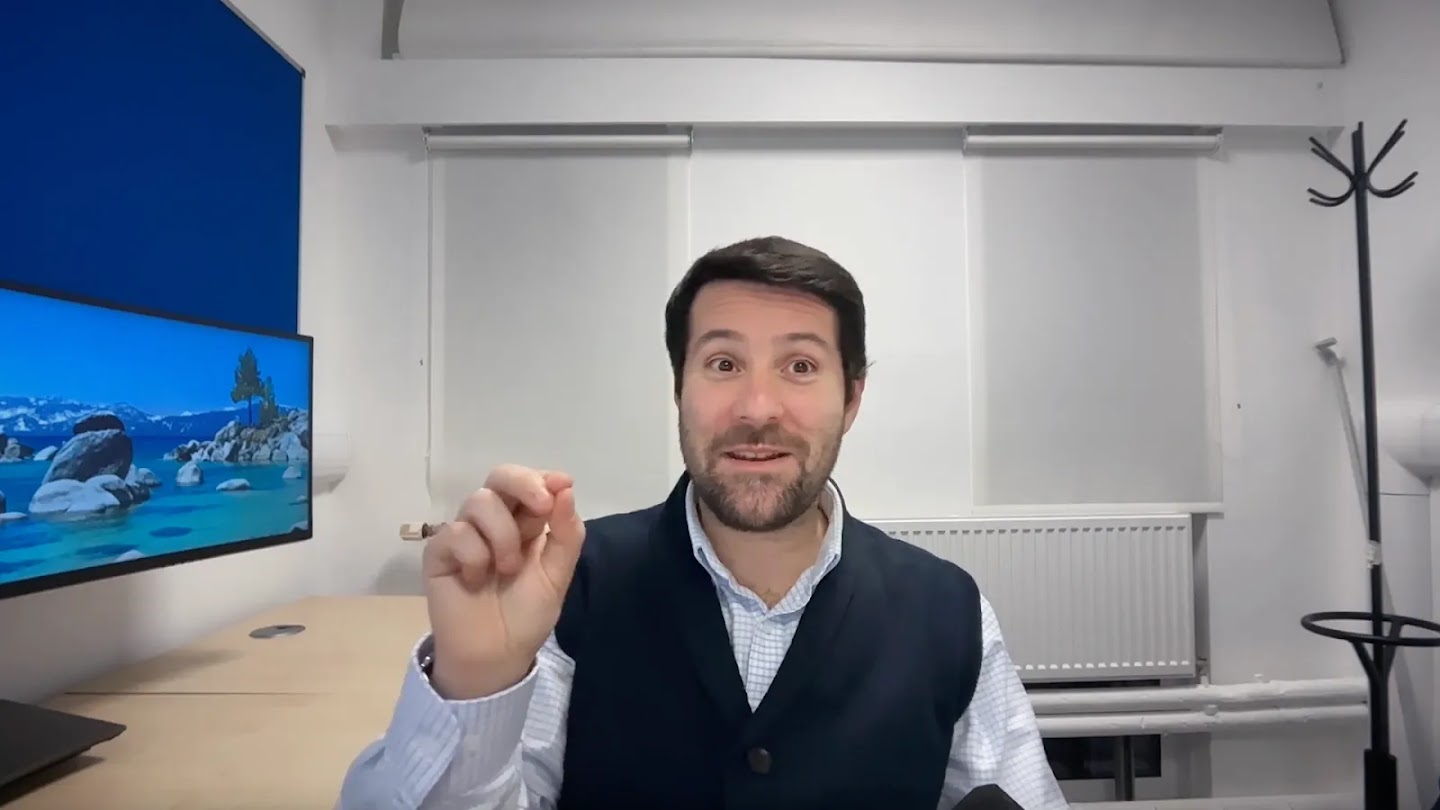 Image 37: A medium shot of a man with dark hair and a beard, smiling and looking directly at the camera. He is wearing a light-colored button-up shirt with a dark blue vest over it. His right hand is raised beside his face, with his thumb and index finger pinched together as if making a gesture or explaining something. The background shows a room with a light blue wall, a computer monitor displaying a natural landscape, and white blinds behind a window.